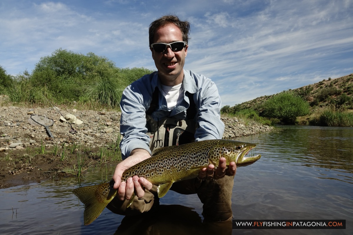 Angler smiling beside the Quemquemtreu stream with rod and net on the gravel bank
